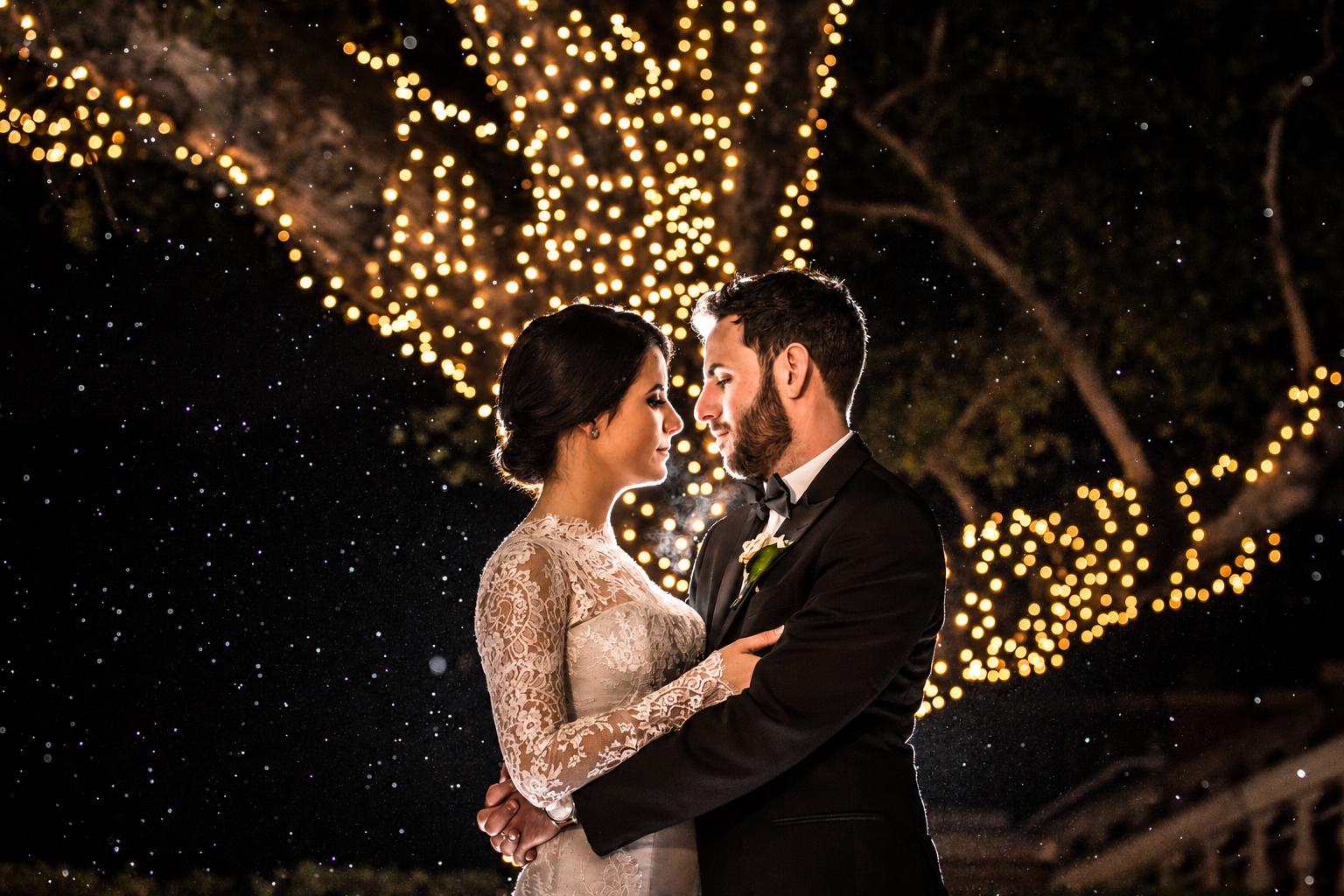 Bride and groom embracing beneath glowing string lights at night, photographed by Shimmy Lautman
